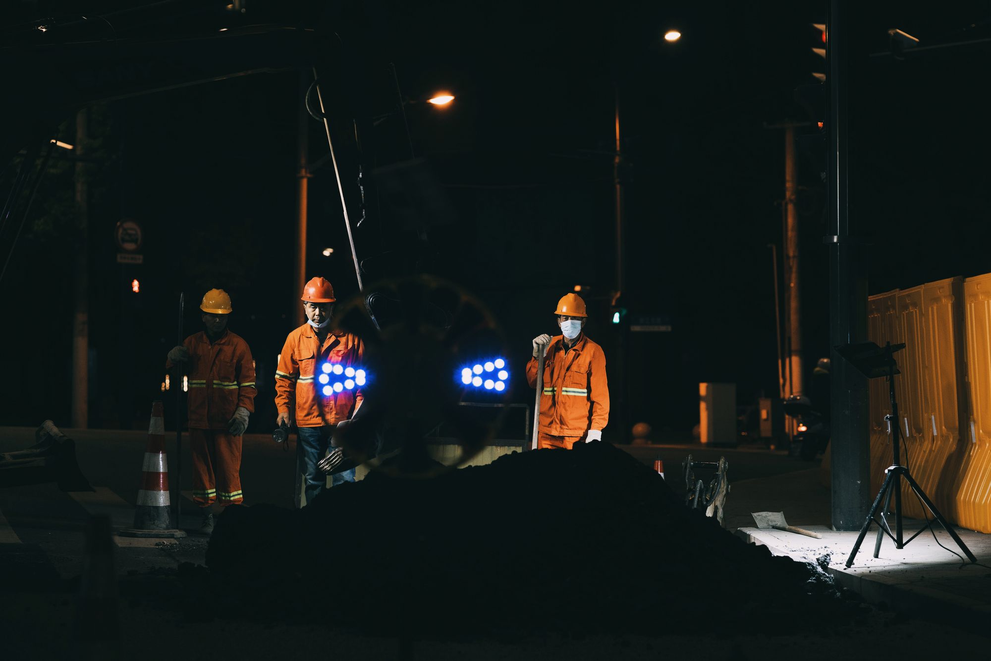 Three workers in orange high-visibility coveralls and hard hats on a night shift roadworks job lit by portable work lights