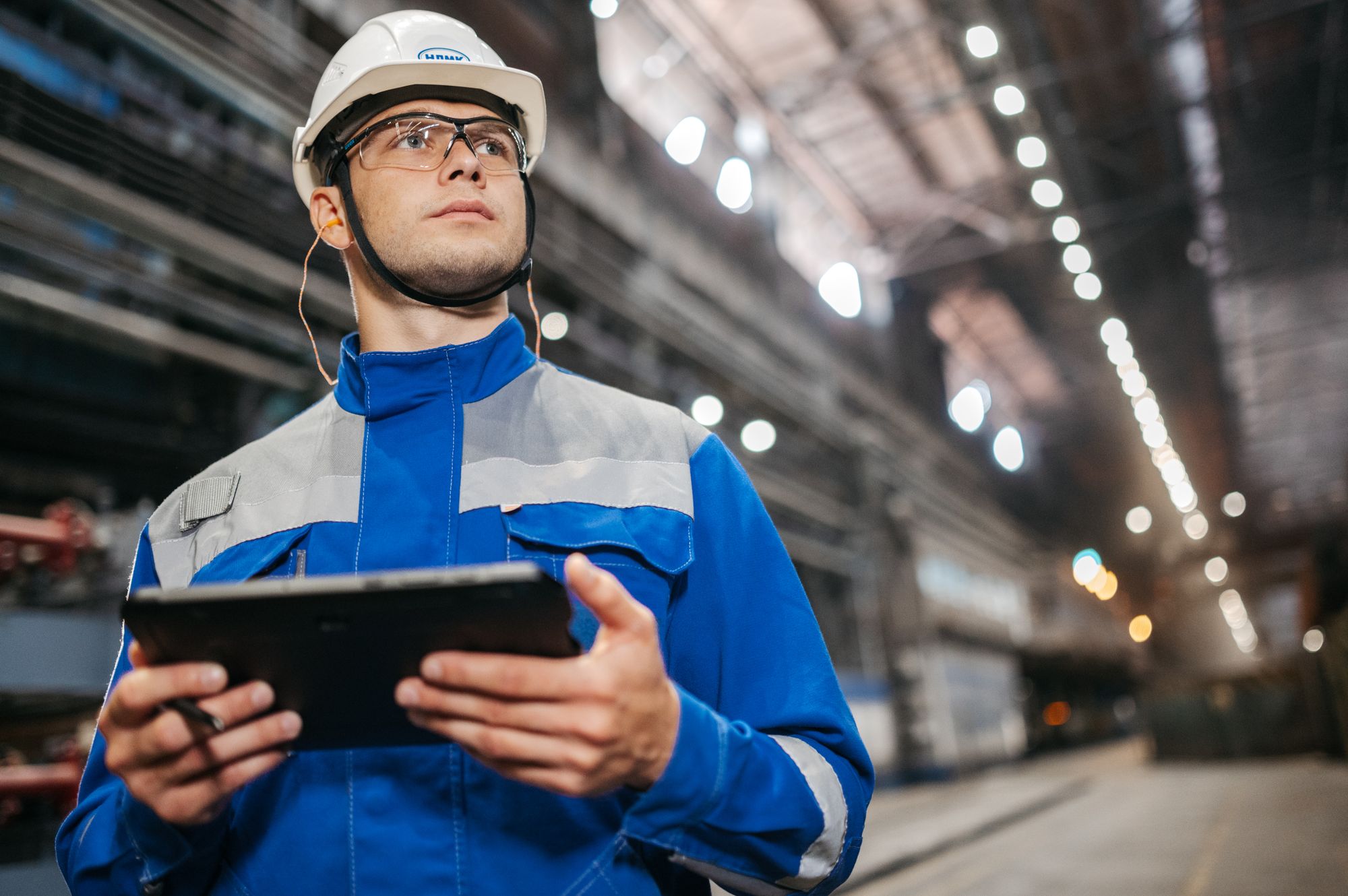 Worker in hard hat and safety glasses holding a tablet on a plant floor, the kind of device used for electronic safety data sheet access