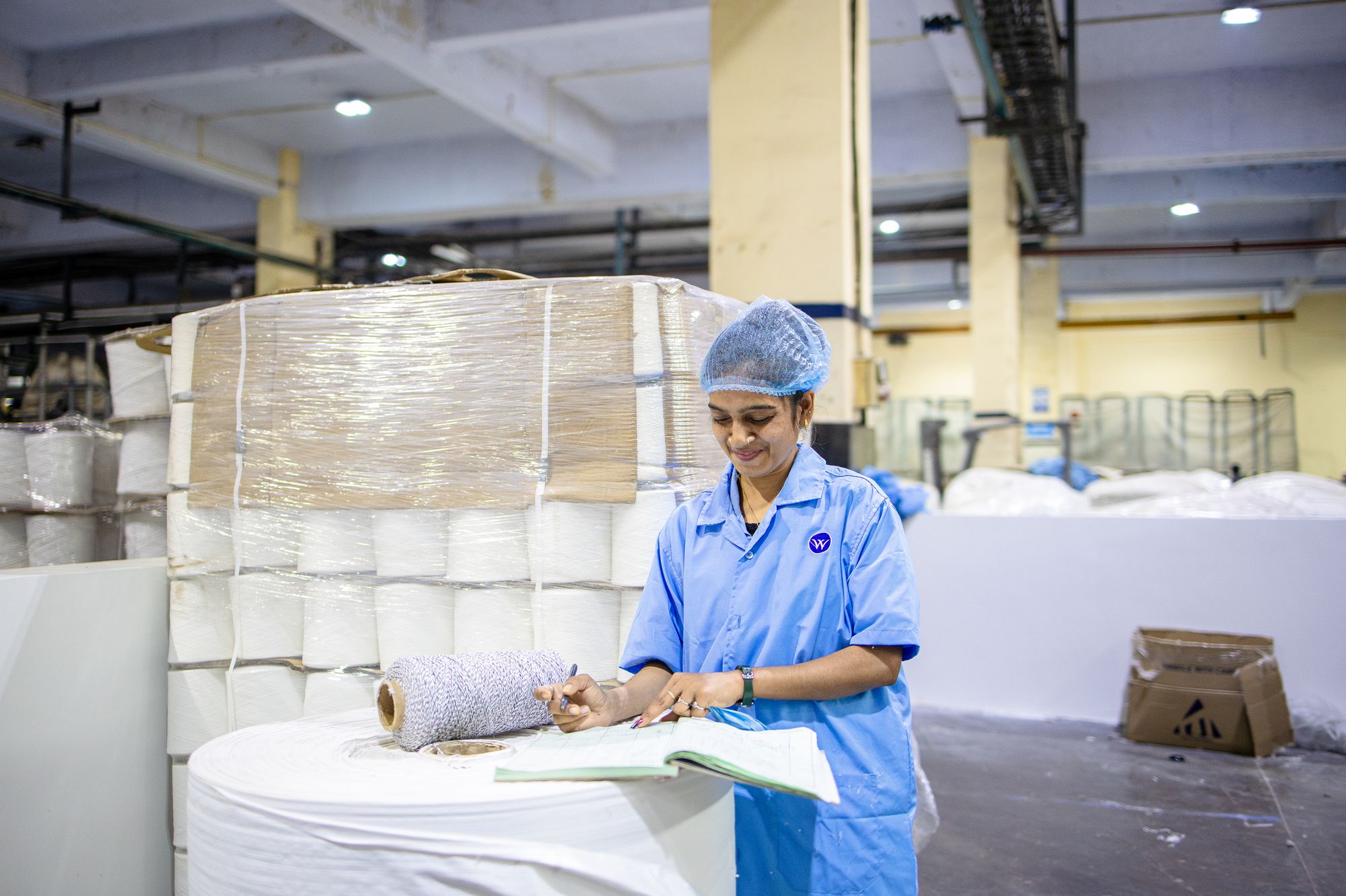 A worker in a blue coat and hairnet writes in an open logbook beside pallets of yarn in a textile plant