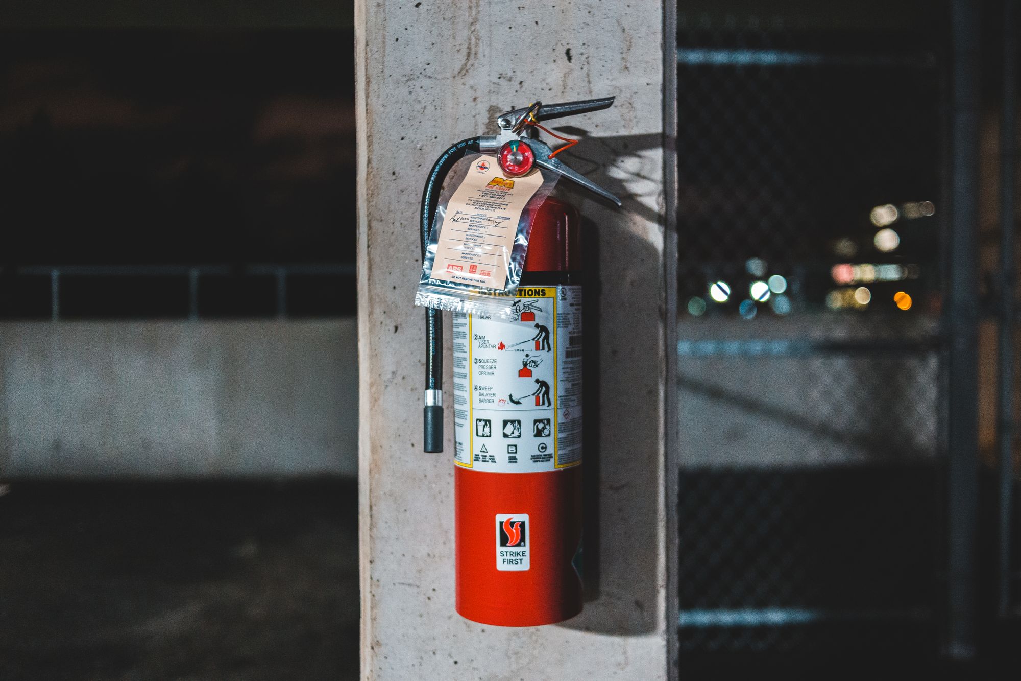 A red ABC dry chemical fire extinguisher mounted on a concrete column with a paper inspection tag hanging from its neck