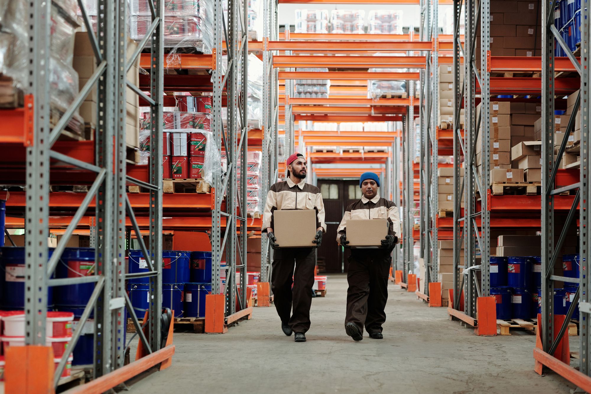 Two warehouse workers carry cardboard cartons by hand along an aisle between tall pallet racks, a manual handling task with repeated lifting and carrying