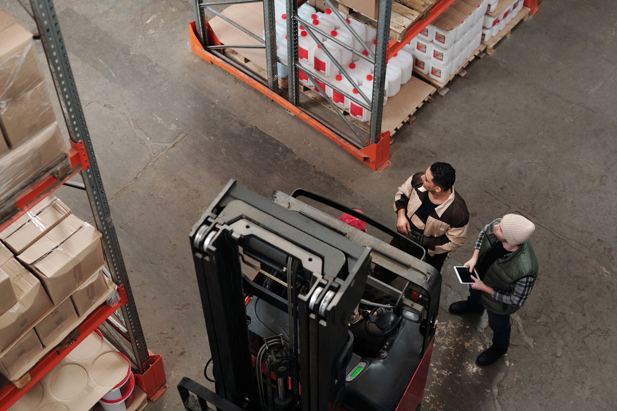 Aerial view of two warehouse workers standing beside a forklift near pallet racking, checking a hazard