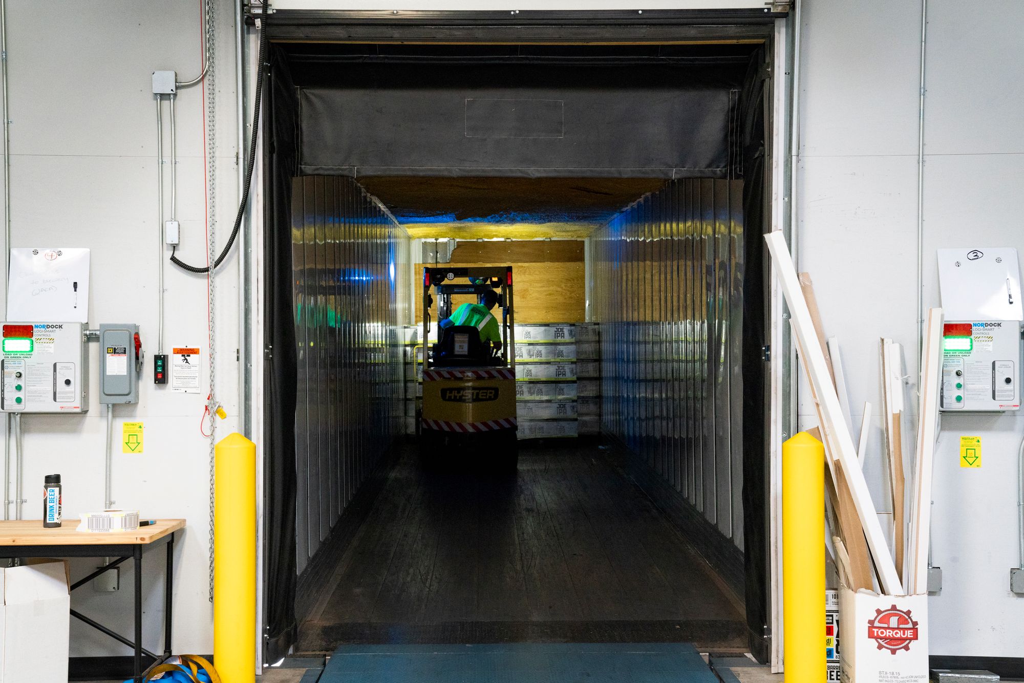 Forklift moving a pallet through a warehouse dock door past yellow protective bollards at a loading bay