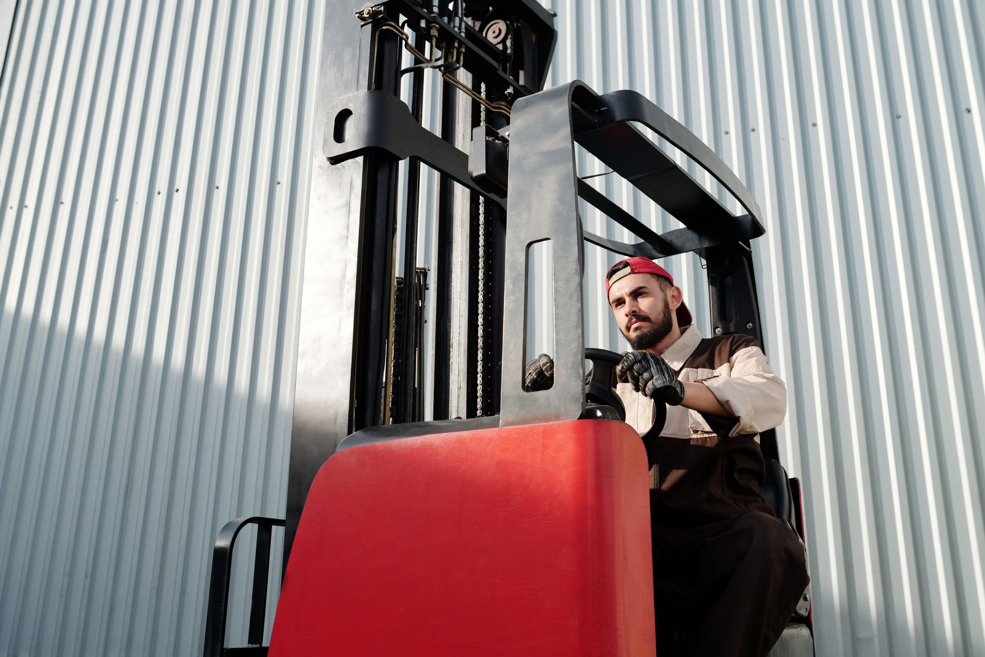 A forklift operator seated in the cab of a red lift truck alongside a warehouse wall, hands on the controls before starting a shift