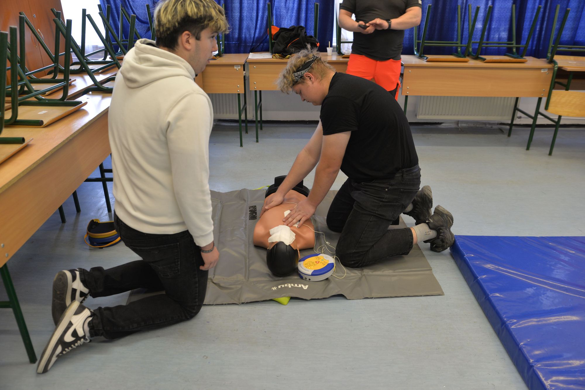 Two trainees kneel over a CPR practice manikin with an AED unit attached during a hands-on first aid training session