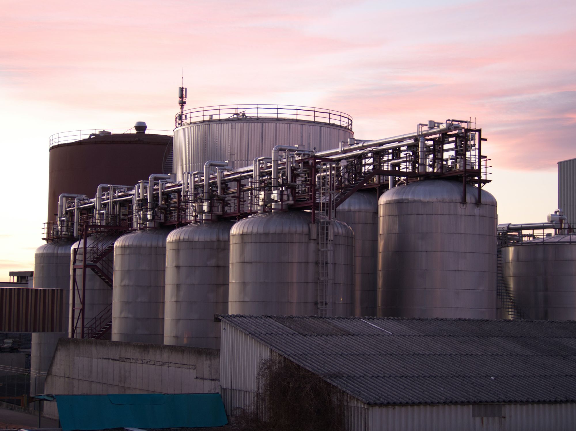 Rows of large process storage tanks connected by overhead pipework at an industrial plant at dusk