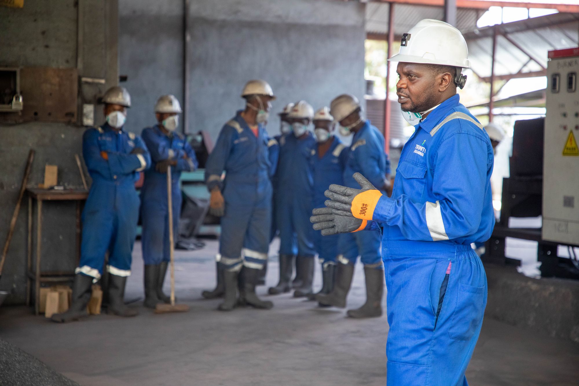 A supervisor in coveralls and a hard hat briefing a group of factory workers wearing hard hats and respirators during a safety training session