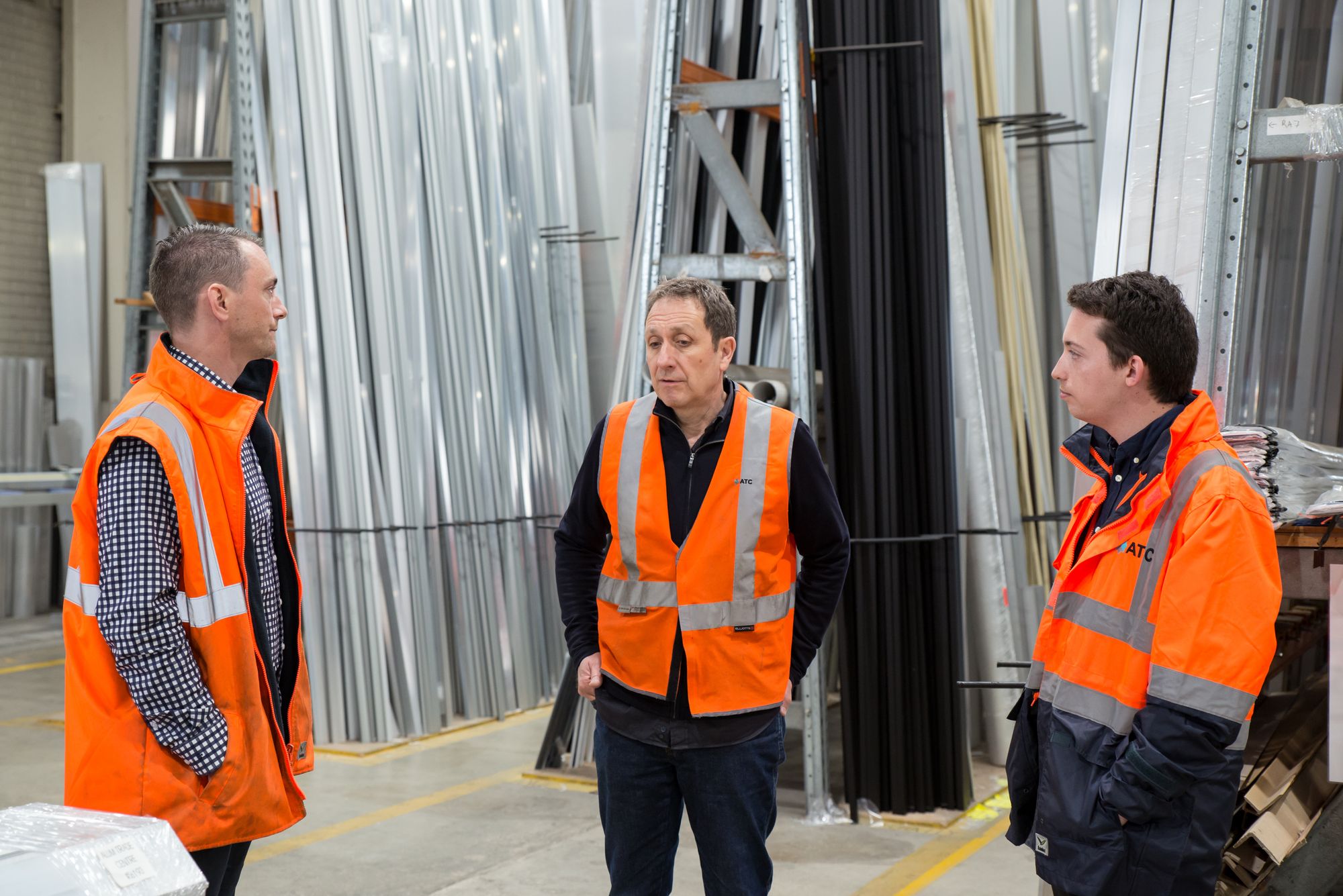 Three warehouse workers in orange hi-vis vests standing among steel racking, talking through a job before it starts