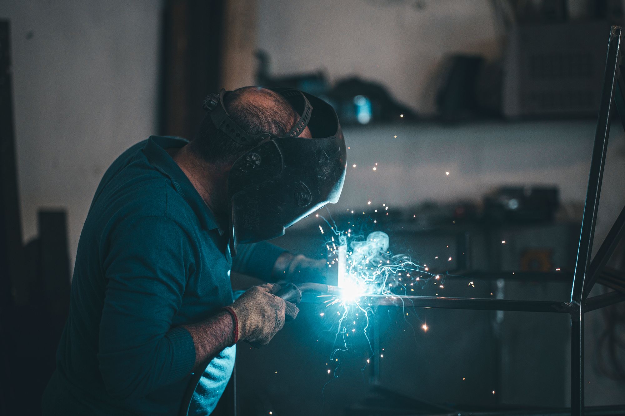 A welder in a helmet and glove strikes an arc on a steel frame in a fabrication shop