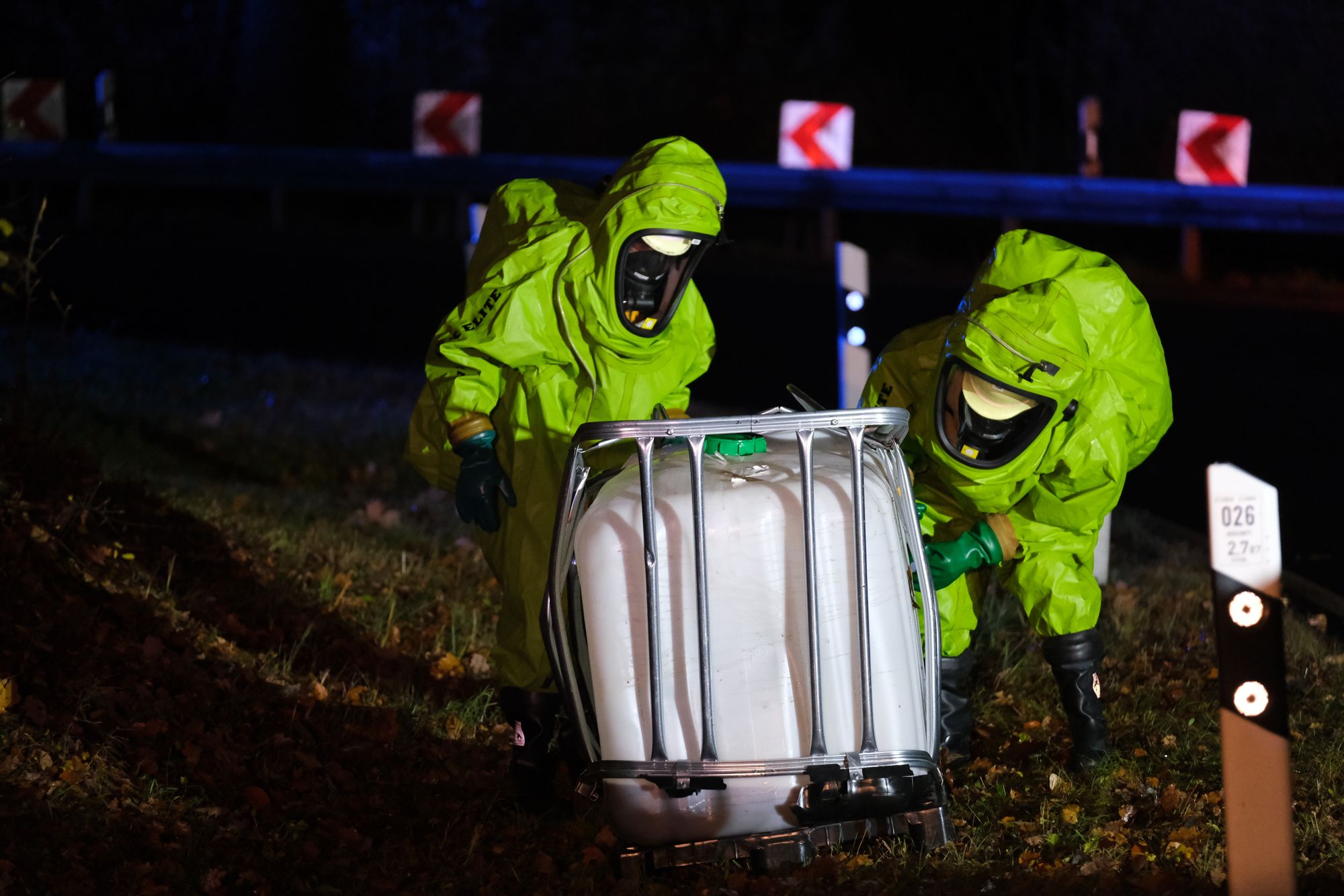 Two hazmat responders in green chemical protective suits steadying a caged IBC tote at night