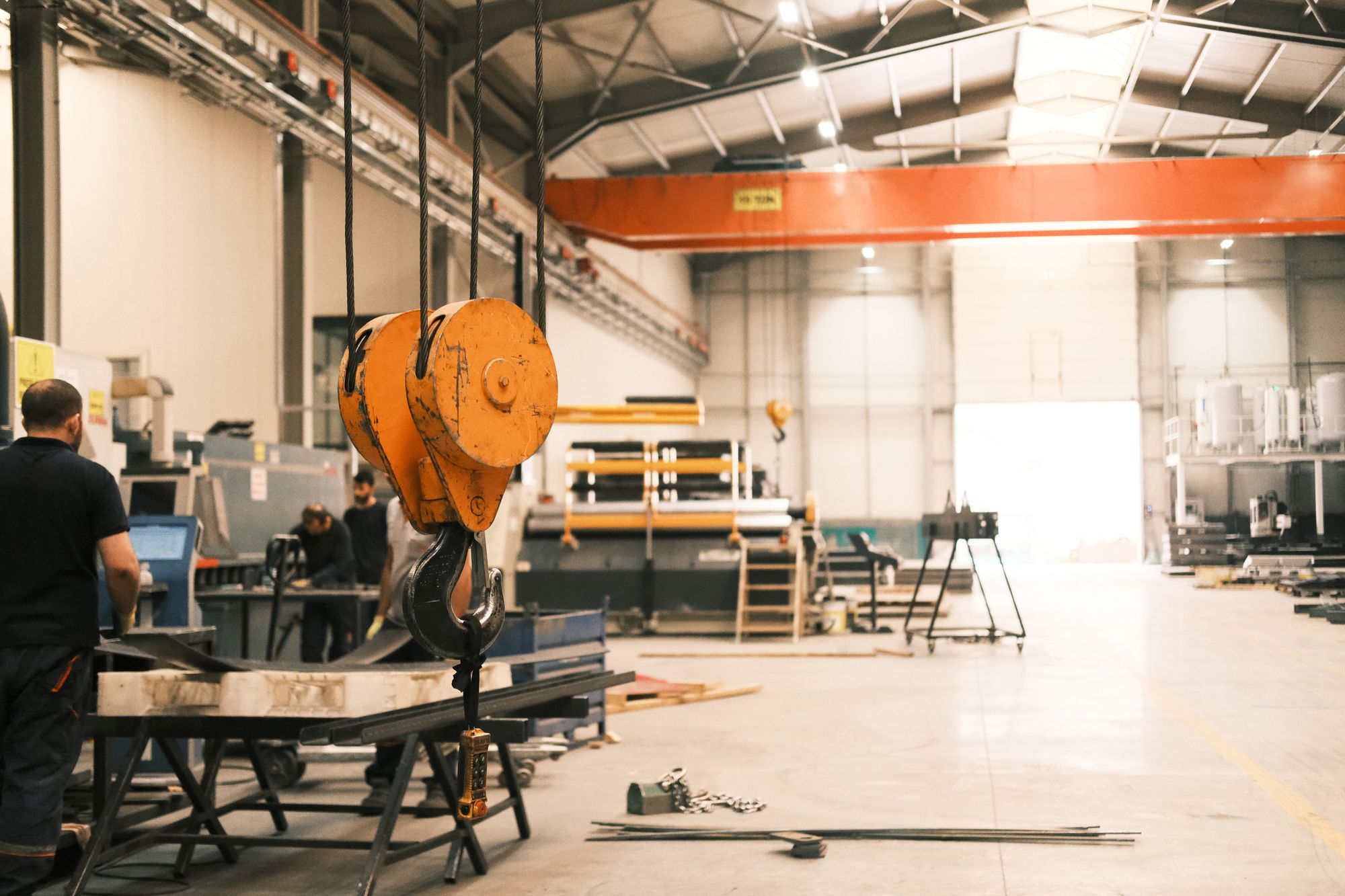Overhead crane hook block hanging on wire rope above a metal fabrication shop floor, with workers at benches in the background