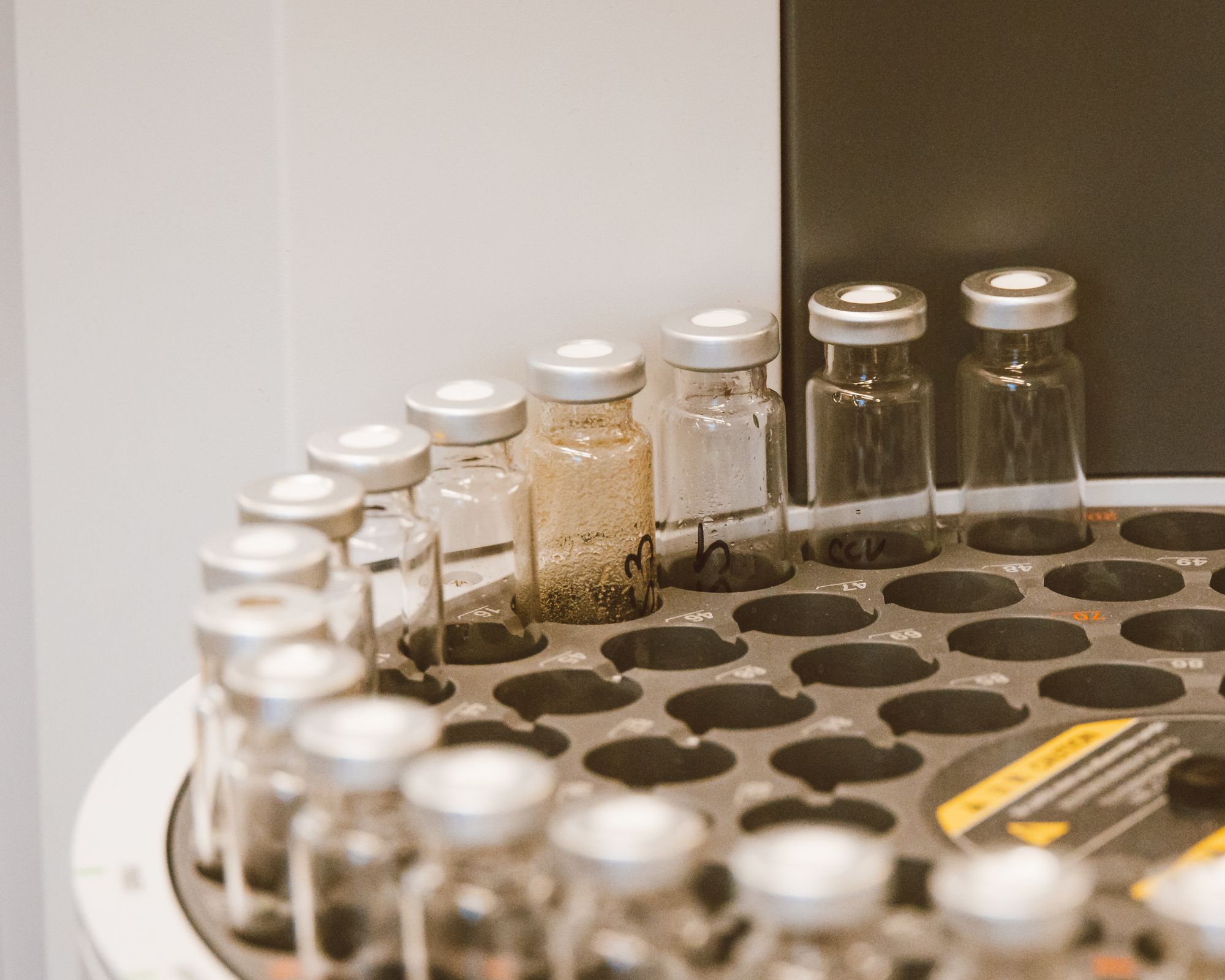 Row of small crimp-sealed glass vials in a laboratory autosampler tray, marked only by hand