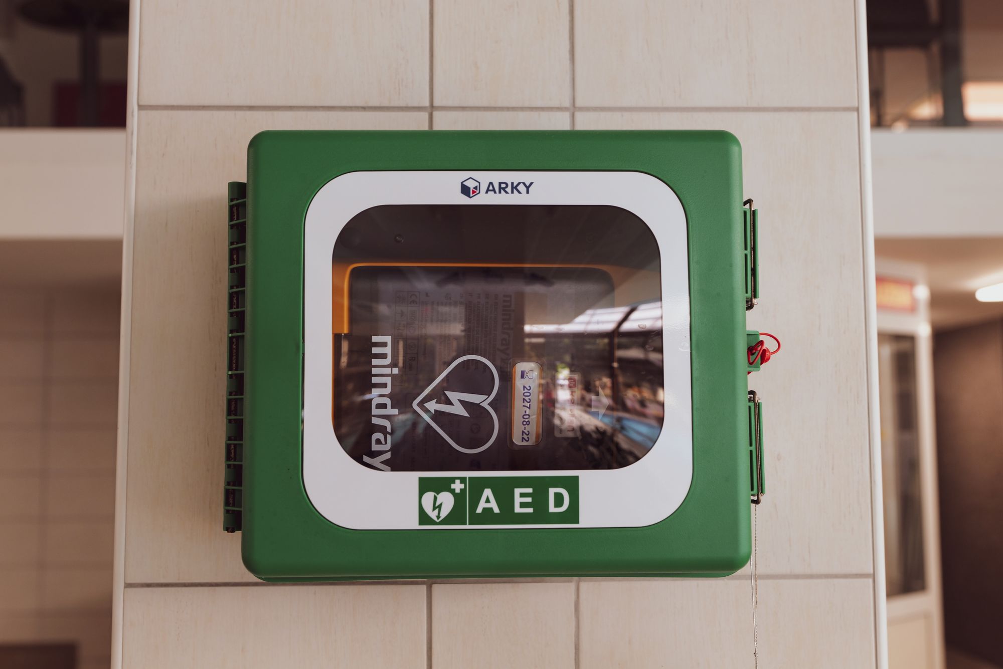 Green wall-mounted AED cabinet with a visible expiration date sticker in a building corridor